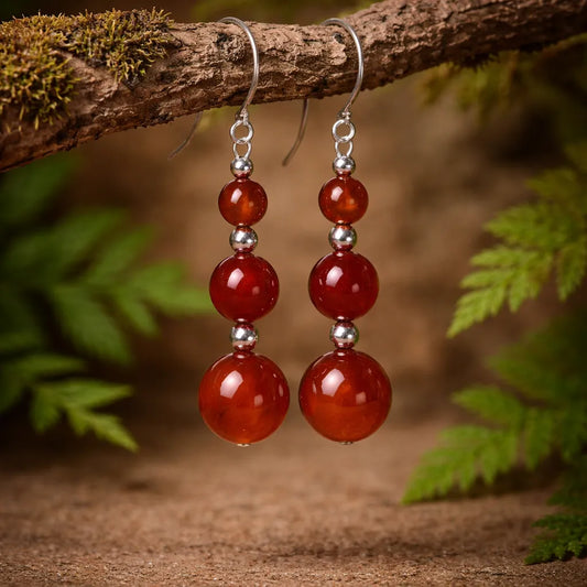 La Boucle d'oreilles en Cornaline de Pierres énergétiques présente trois perles de cornaline rouge-orange brillantes, séparées par des entretoises en argent, suspendues à des crochets. Elle est présentée sur une branche avec de la verdure et de la mousse en arrière-plan.